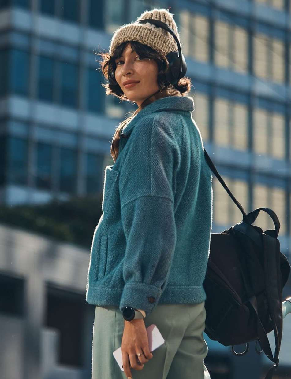 Woman in a teal jacket, beige knit hat, and headphones outdoors in an urban setting, holding a phone, with a backpack. Medium shot.