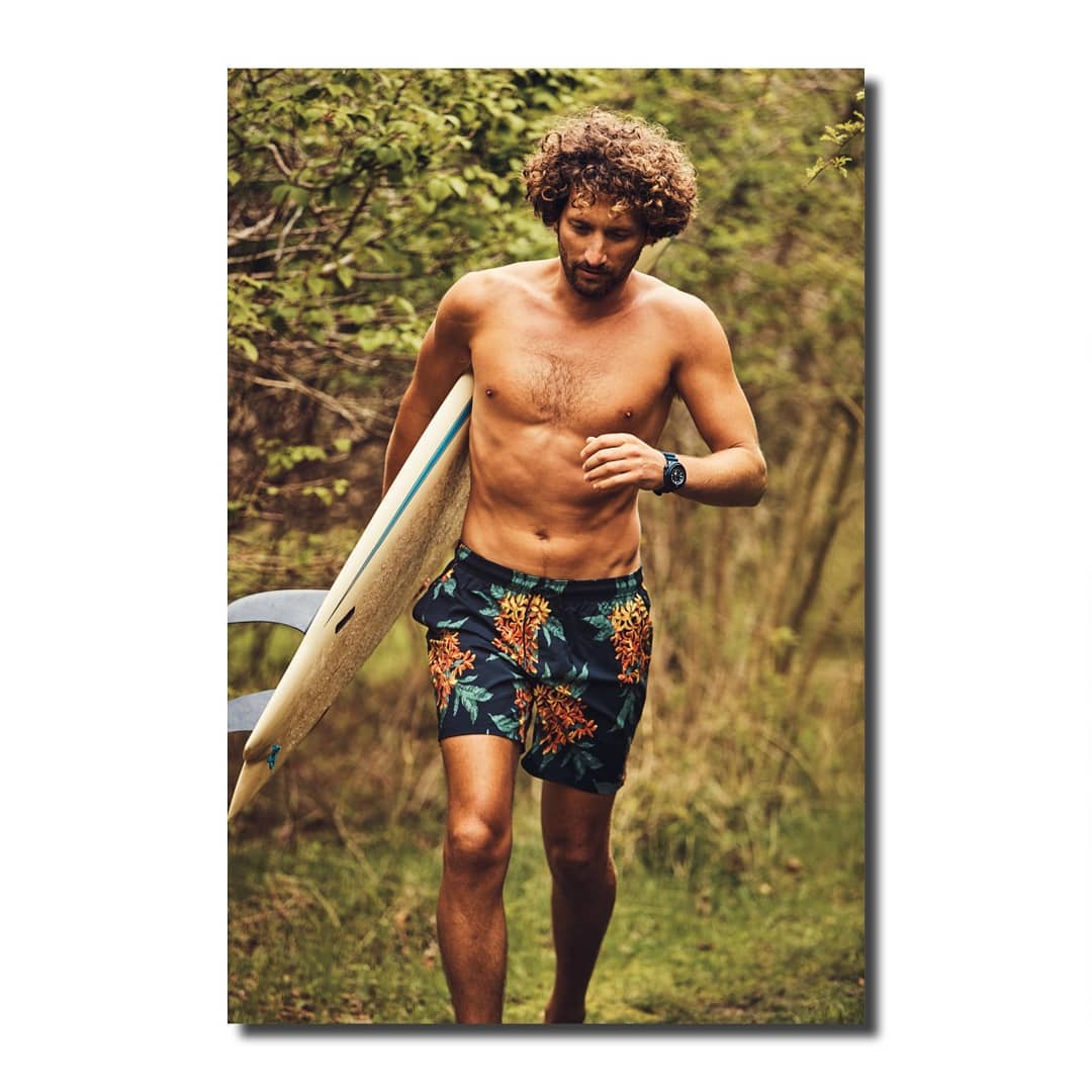 Man walking shirtless with surfboard on a nature trail, wearing patterned shorts and a watch, surrounded by green foliage.
