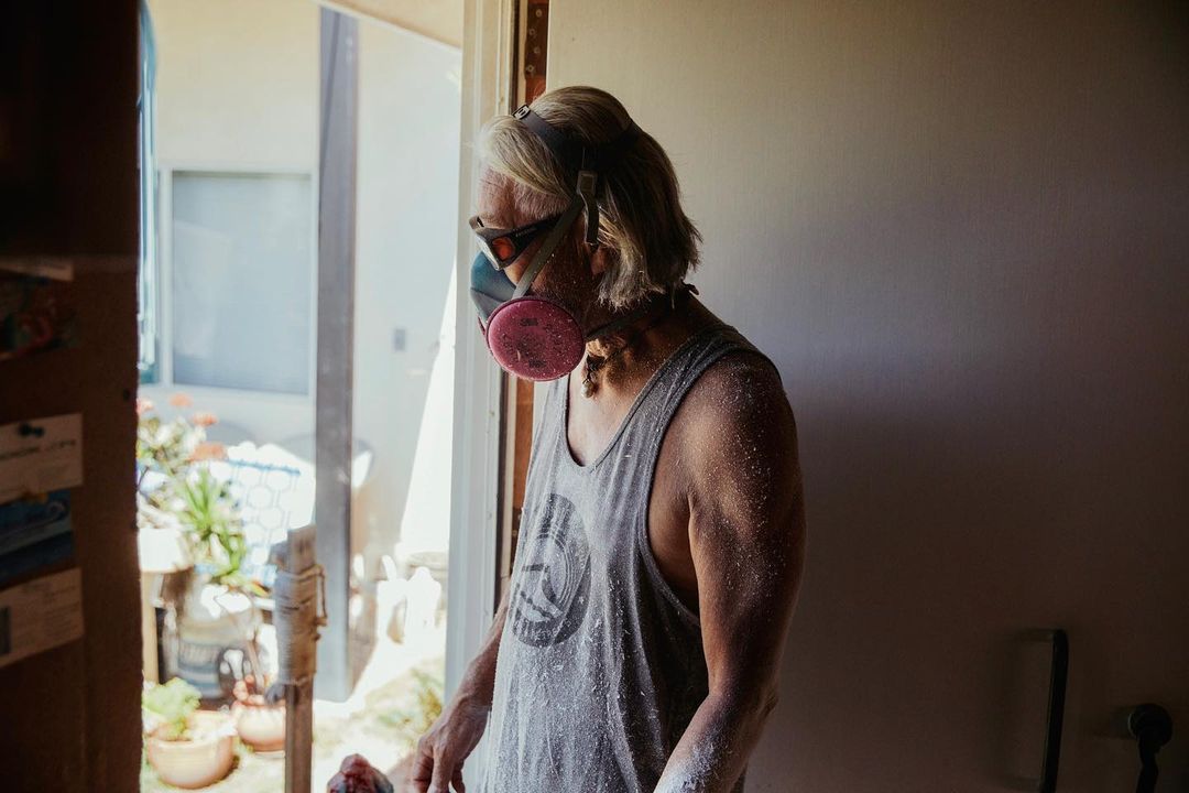 An adult wearing a gray tank top and a pink respirator mask stands indoors near a doorway with an outdoor view and dappled lighting.