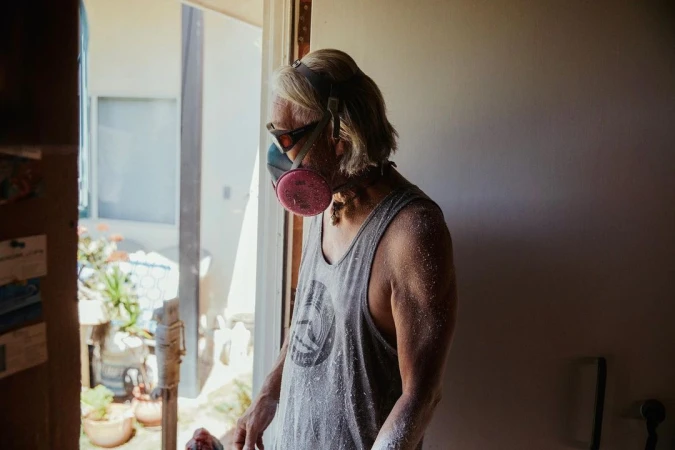 An adult wearing a gray tank top and a pink respirator mask stands indoors near a doorway with an outdoor view and dappled lighting.