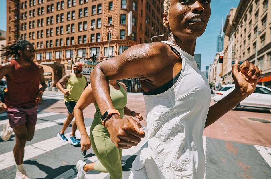 A group of four athletic people running across a city street crosswalk on a sunny day wearing activewear and fitness trackers on their wrists.