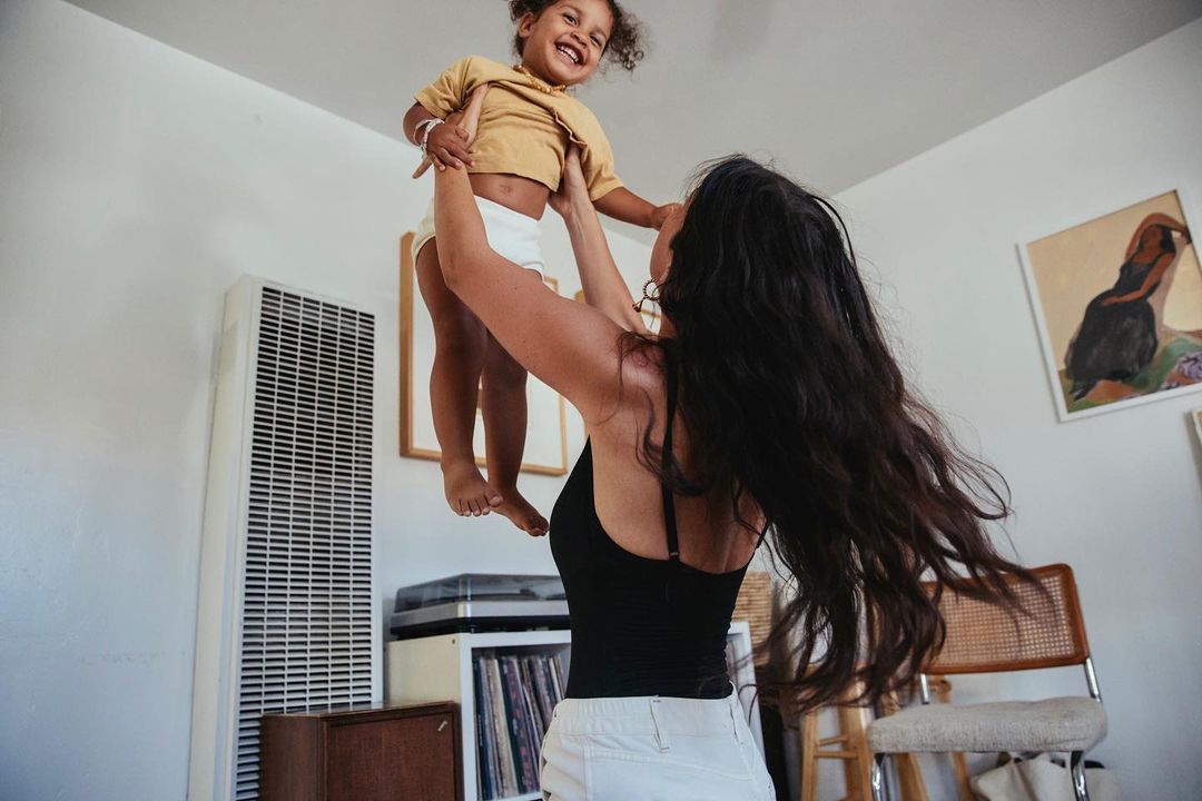 A happy child is lifted high into the air by an adult in an indoor setting with white walls and casual clothing, creating a joyful scene.