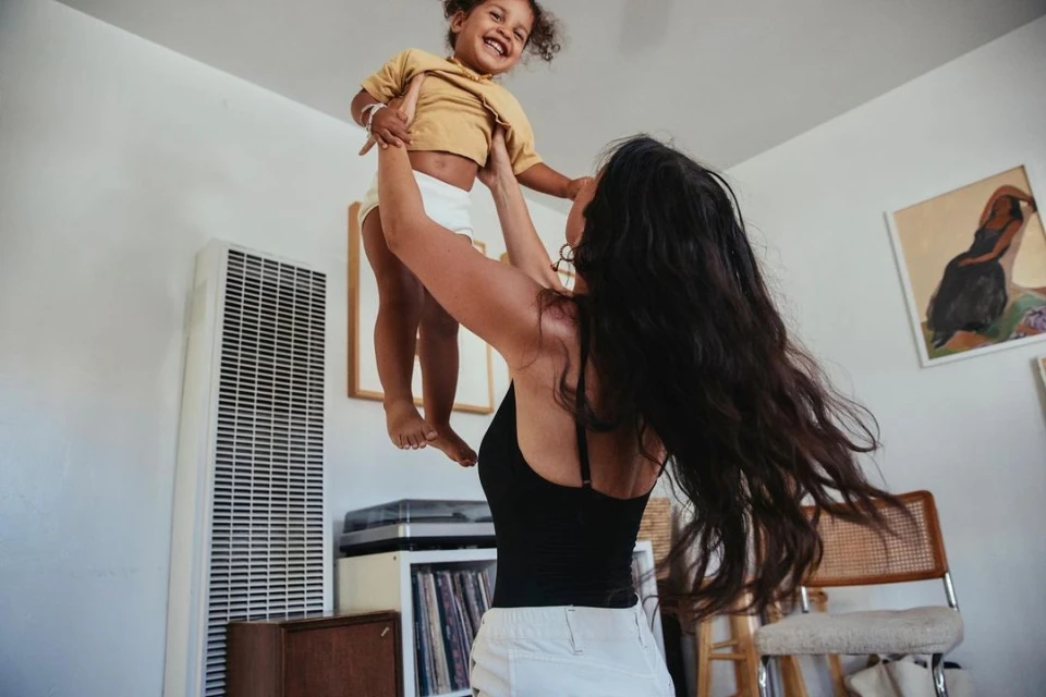 A happy child is lifted high into the air by an adult in an indoor setting with white walls and casual clothing, creating a joyful scene.