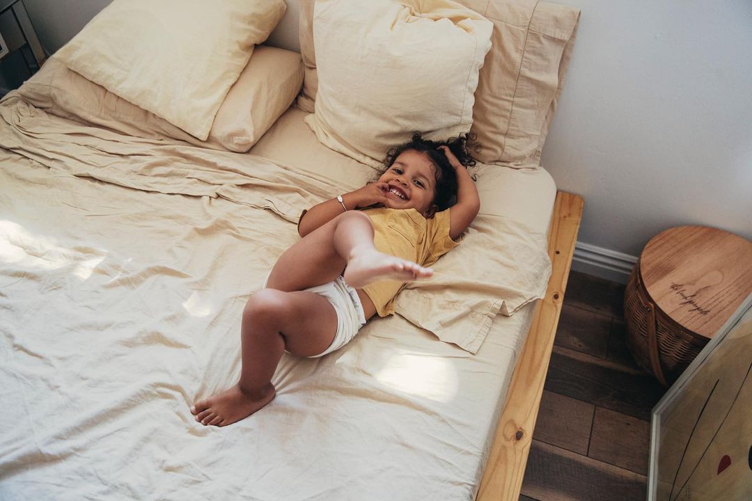 Happy toddler lying on a bed in a yellow t-shirt and white diaper, smiling and looking up at the camera in an indoor setting.