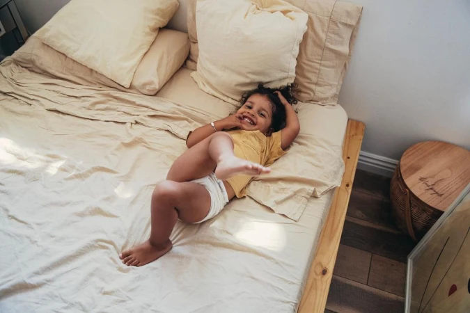 Happy toddler lying on a bed in a yellow t-shirt and white diaper, smiling and looking up at the camera in an indoor setting.