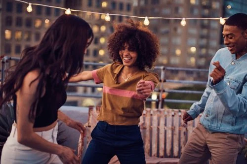 Three young friends dancing outdoors on a rooftop at dusk under string lights, captured by Jeff Vallee in New York.