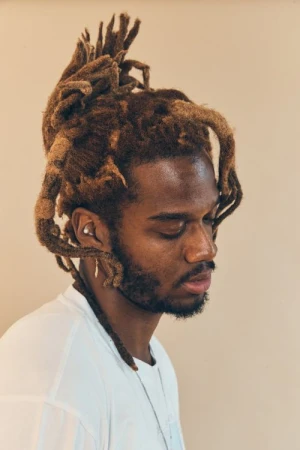 Portrait of a black man with locs styled in a high half-up bun wearing a white t-shirt against a plain neutral wall.