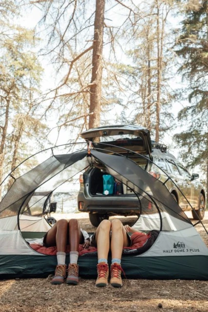 Two campers relax inside an REI Half Dome 3 Plus tent outdoors near a parked SUV in a wooded area, showcasing a camping lifestyle.