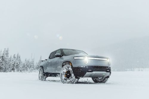 A gray Rivian pickup truck drives through a snowy field with a forest backdrop, under an overcast sky, showcasing its electric vehicle capabilities.