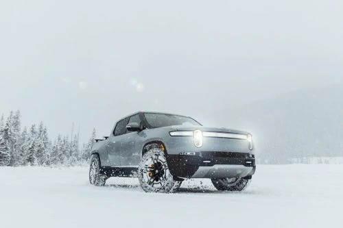A gray Rivian pickup truck drives through a snowy field with a forest backdrop, under an overcast sky, showcasing its electric vehicle capabilities.