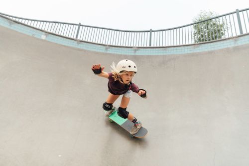 Young child on a skateboard wearing safety equipment in a skatepark, practicing the sport, with a grey concrete ramp background.