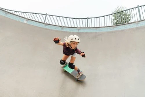 Young child on a skateboard wearing safety equipment in a skatepark, practicing the sport, with a grey concrete ramp background.