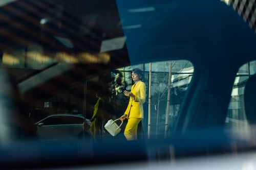 Woman in yellow suit holding a white bag standing in front of a modern building with glass facade as seen through a car window.