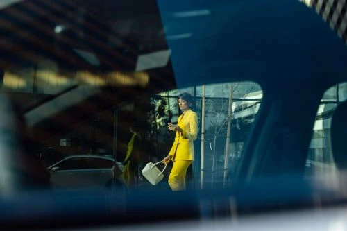 Woman in yellow suit holding a white bag standing in front of a modern building with glass facade as seen through a car window.
