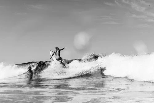 Black and white photo of a surfer riding a wave on a surfboard under a bright sky. The wave is breaking with white foamy water.