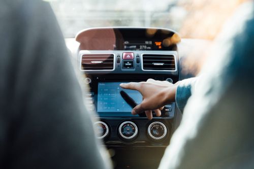 Interior car view featuring an adult's hand interacting with the infotainment screen, dashboard, and steering wheel with a blurred passenger.