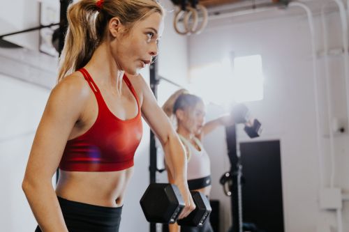 Two women are exercising indoors, lifting dumbbells in a bright space with gym equipment. One woman wears red, the other wears white.