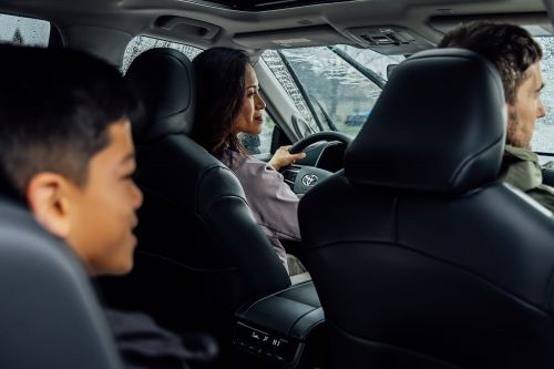 Family in a car on a rainy day with a woman driving. The backseats are dark leather and the windshield wipers are on.