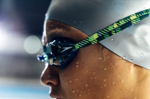 Close-up shot of an adult female swimmer wearing a white swim cap and blue goggles, with water droplets on her face.