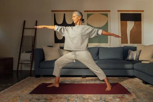 An older adult female doing yoga in an indoor setting, wearing athleisure clothing, with a couch and artwork visible in the background.