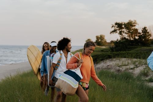 A group of friends walk along a grassy beach path with a surfboard and tote bag near the ocean at dusk, enjoying a beach outing.