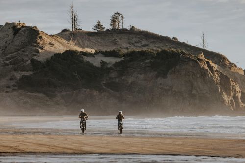 Two motorcyclists ride across a sandy beach with a large rocky headland in the background on a clear, sunny day.