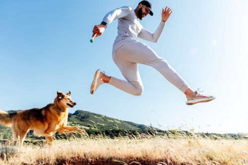 Athletic man jumps high in the air, running and playing with his dog in a grassy field with a mountain in the background, sunny day.