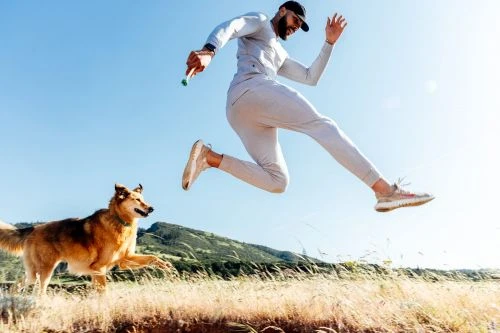 Athletic man jumps high in the air, running and playing with his dog in a grassy field with a mountain in the background, sunny day.