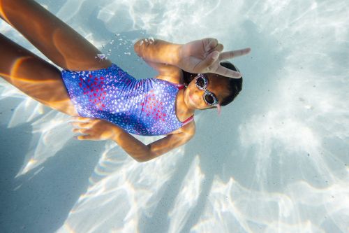 A young girl swims underwater in a blue swimsuit with a peace sign, sunlight shimmering through the water in an outdoor pool.