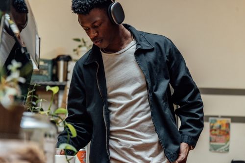 Black man wearing over-ear headphones and a dark zip-up jacket over a white t-shirt in a domestic interior setting.