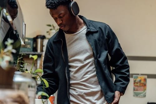 Black man wearing over-ear headphones and a dark zip-up jacket over a white t-shirt in a domestic interior setting.