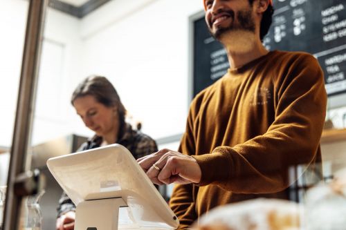 Man using POS system with woman in cafe. The man is wearing a brown shirt and a beanie. Interior setting with natural light.