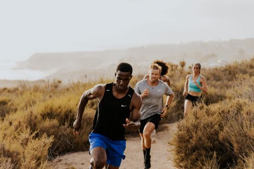 A group of four athletic people running uphill on a dirt path outdoors, with an ocean view in the background during daylight.