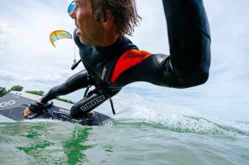 A male kitesurfer in a wetsuit rides a board on the ocean, with a kite in the background against a cloudy sky.