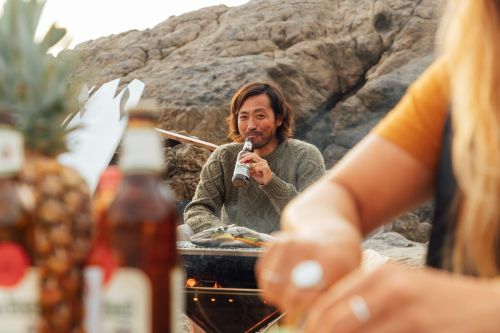 An Asian man drinks a beer on a rocky beach while grilling food, with a woman and pineapples visible in the foreground.