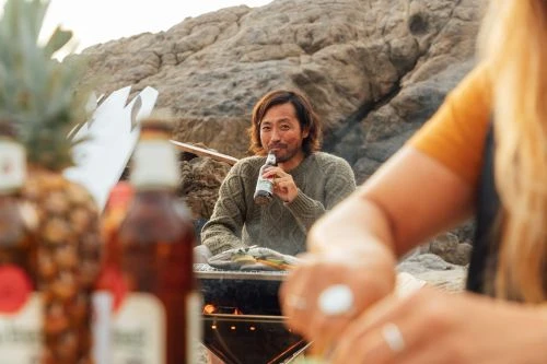 An Asian man drinks a beer on a rocky beach while grilling food, with a woman and pineapples visible in the foreground.