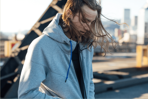 Young man with long hair wearing a grey zip hoodie outdoors with city skyline background