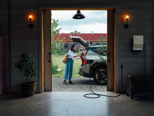 Woman next to electric vehicle plugged in at home. Car is parked near open garage door overlooking trees and a red-roofed building.