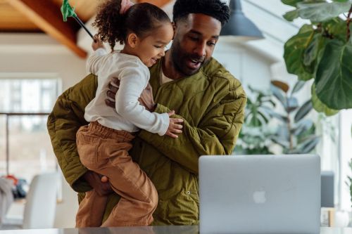 Father holds young daughter as they both look at a laptop in a brightly lit home interior. The daughter holds a toy wand.