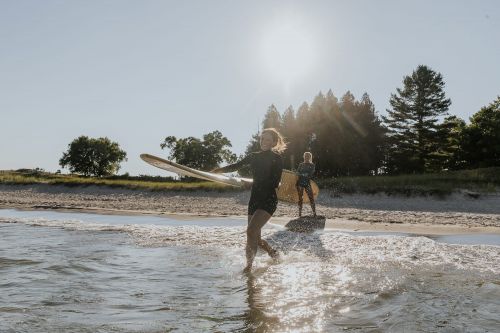 Two women in wetsuits run with surfboards in shallow ocean water near a sandy beach with trees on a sunny day.