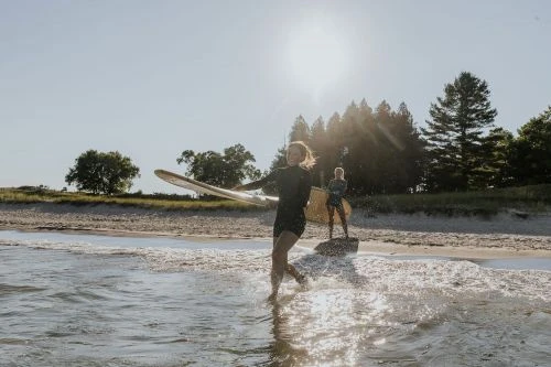 Two women in wetsuits run with surfboards in shallow ocean water near a sandy beach with trees on a sunny day.