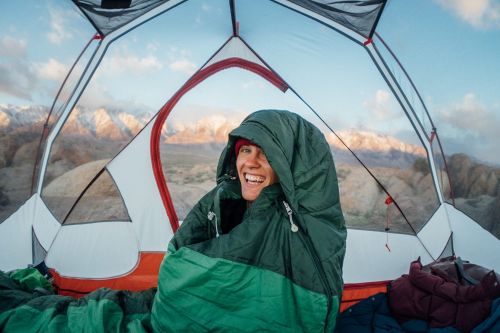 Smiling adult in sleeping bag inside tent; mountain landscape background. Camping and outdoor adventure photography.
