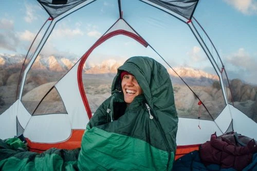 Smiling adult in sleeping bag inside tent; mountain landscape background. Camping and outdoor adventure photography.