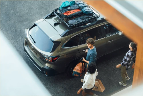 Family loads up a brown SUV with roof cargo carrier in front of a building; man opens car door with bags and shopping tote.