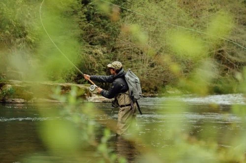 Man fly fishing in a river surrounded by lush green trees. Wearing a hat and backpack, he casts a line into the water.