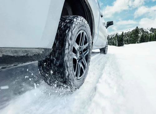 Car tire driving through fresh snow with snow-covered landscape and forest in background under blue sky, winter conditions. 
