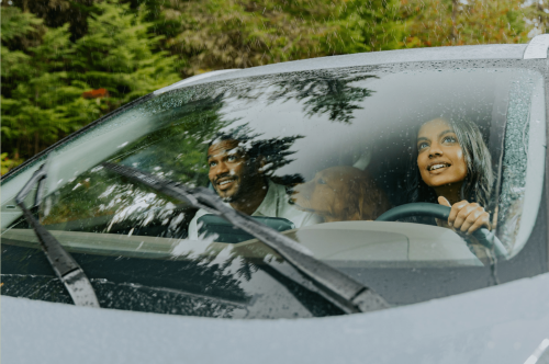 A happy couple with their dog, driving in a car on a rainy day with green trees in the background, seen through the windshield.