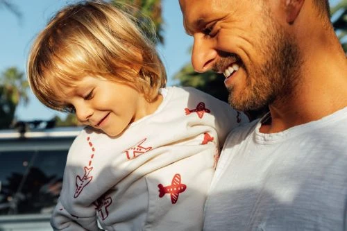 A blond-haired child is being held by an adult male in outdoor setting. The child wears a sweatshirt with airplane patterns.
