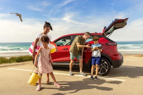 Family of four at the beach unpacks a red car, with the ocean and blue sky in the background, creating a scene of leisure and travel.