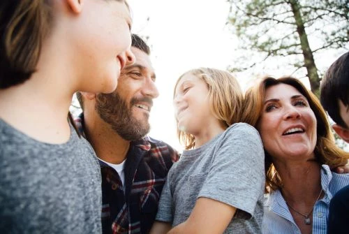 Low angle portrait of a smiling family of five including two children and parents posing together outdoors in dappled sunlight.